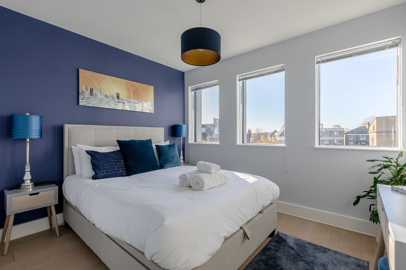 A regular size bedroom with a dark navy accent colour wall, 3 windows, a king size bed with white linens and accent colour decorative cushions.