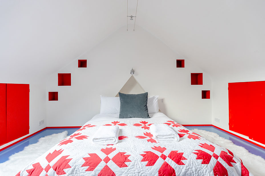 An attic bedroom with white walls, bright red used as an accent colour, bed cover has patterns with same red and cushions used for decoration, shot for property photography.
