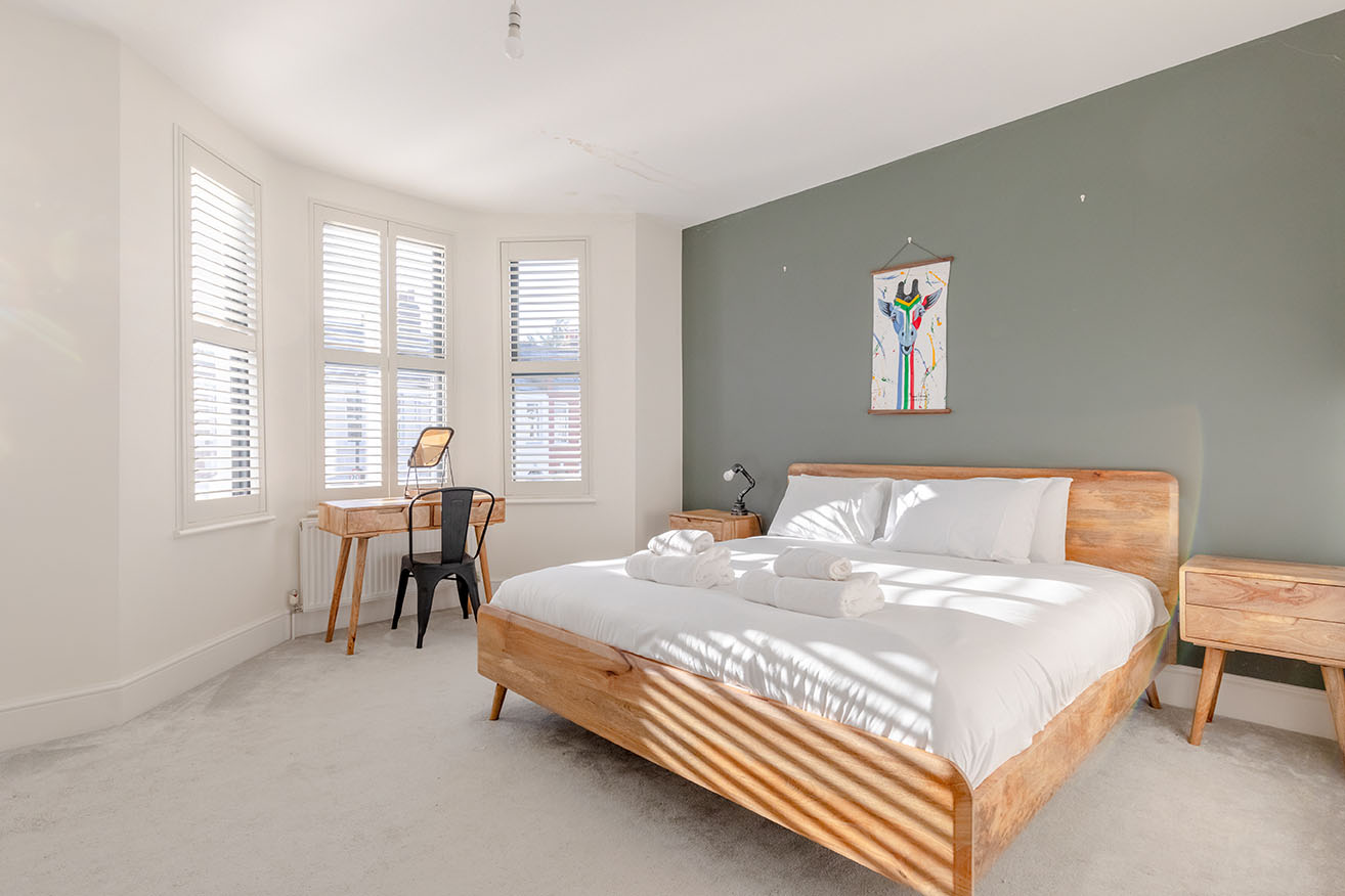 A large bedroom decorated with neutral colours, olive green accent wall, natural wood bedframe and white linens photographed with cross angle for property photography.