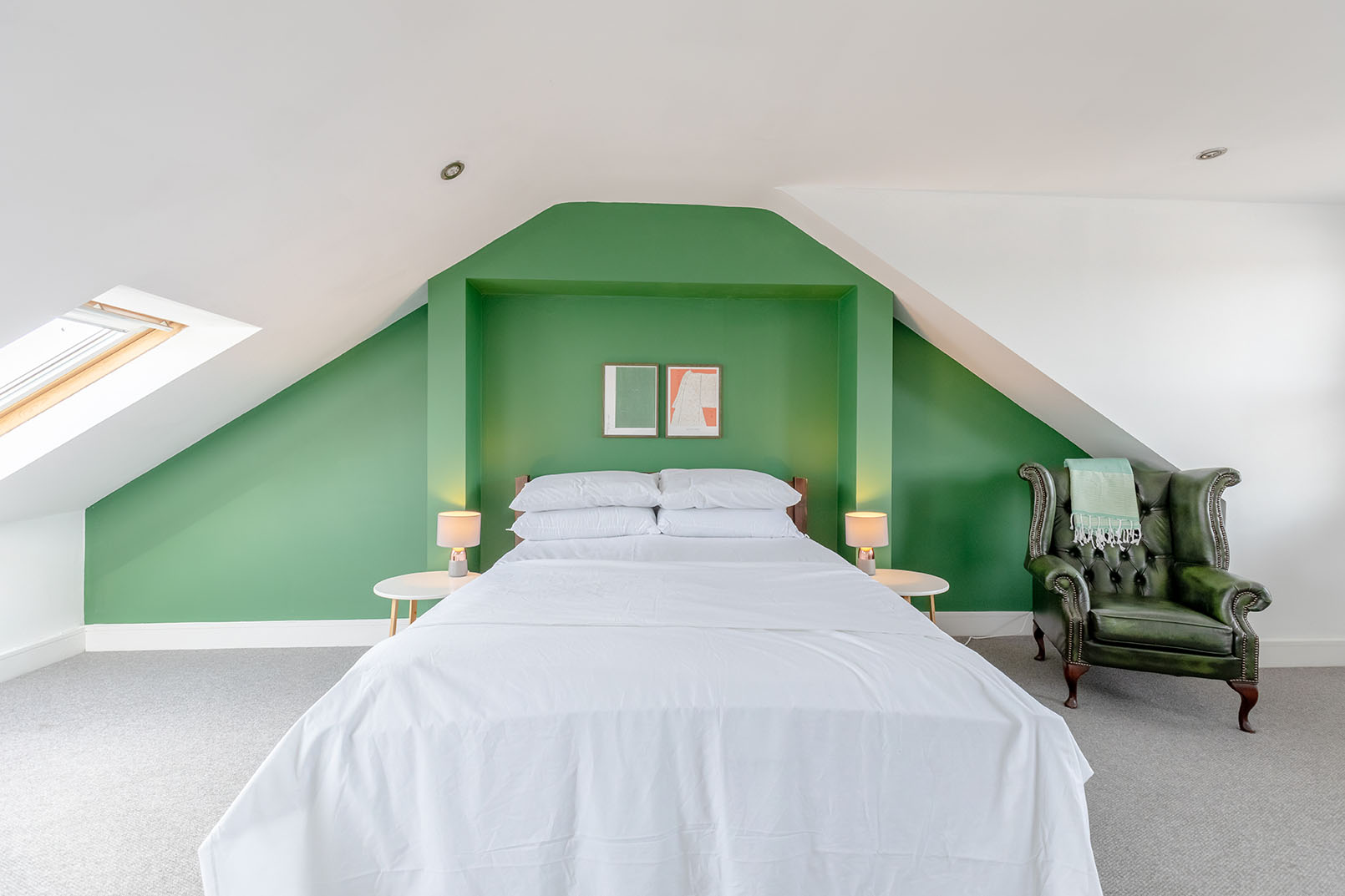 A bright attic bedroom with an accent bright green wall, white linens and a nice green armchair next to the bed, direct angle from the end of the bed.