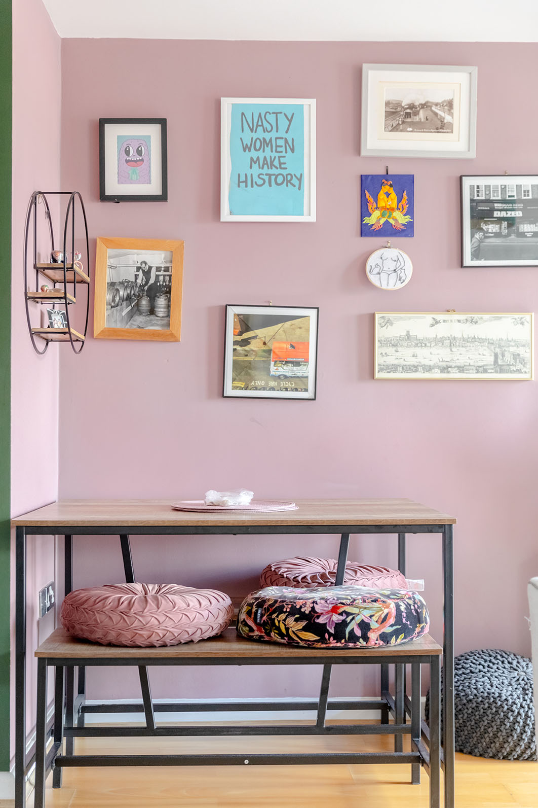 a light pink coloured wall of a dining area, with a wooden and black metal legged dining table, a bench and coloured cushions on the bench, lots of posters hanging on the wall.