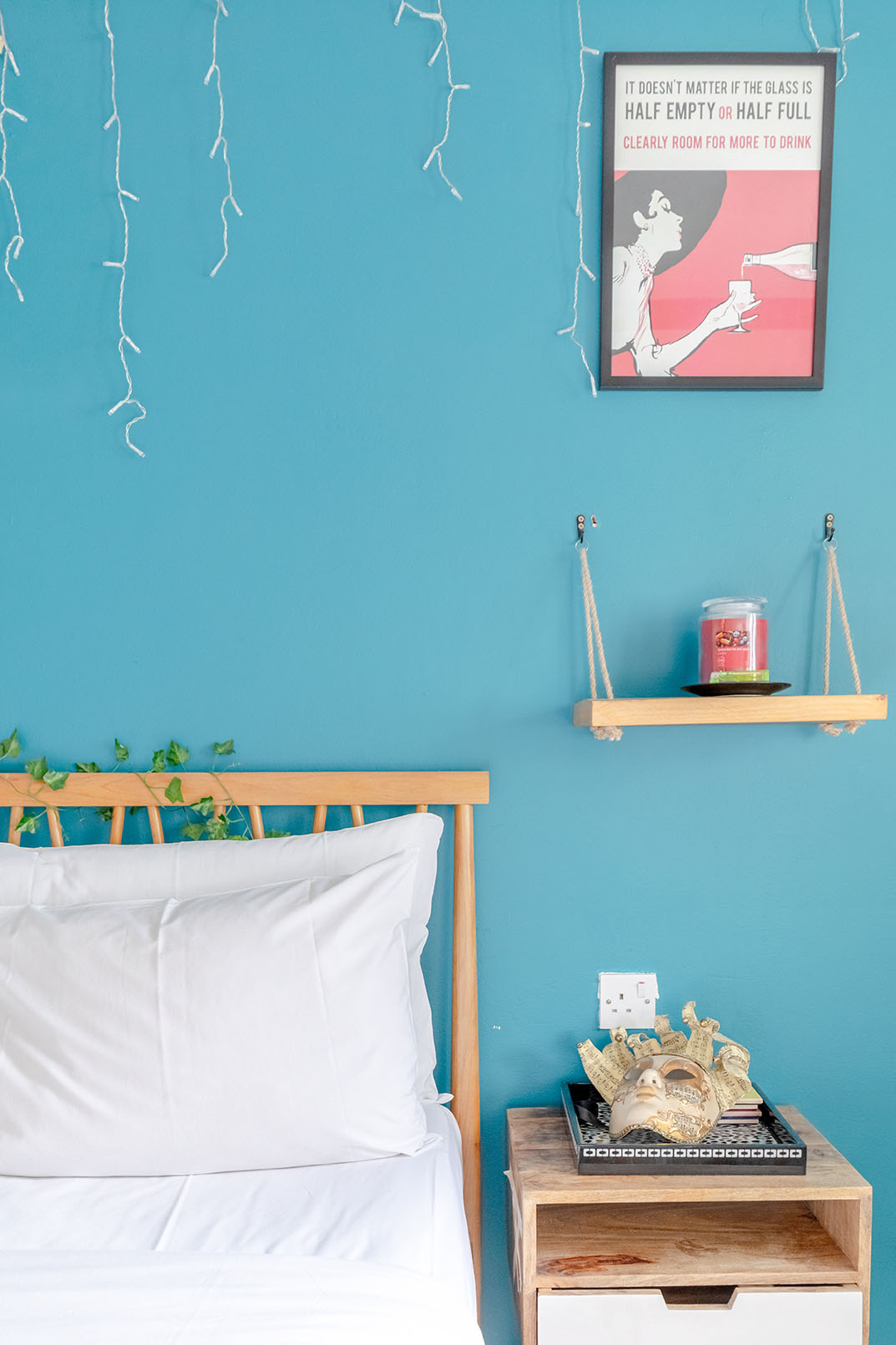 A turquoise coloured wall of a bedroom, the corner of the bed is captured, it's wooden, the bed has white linens, there's a hanging wooden shelf, a poster and a wooden natural bedside table, taken for property photography as a detail.