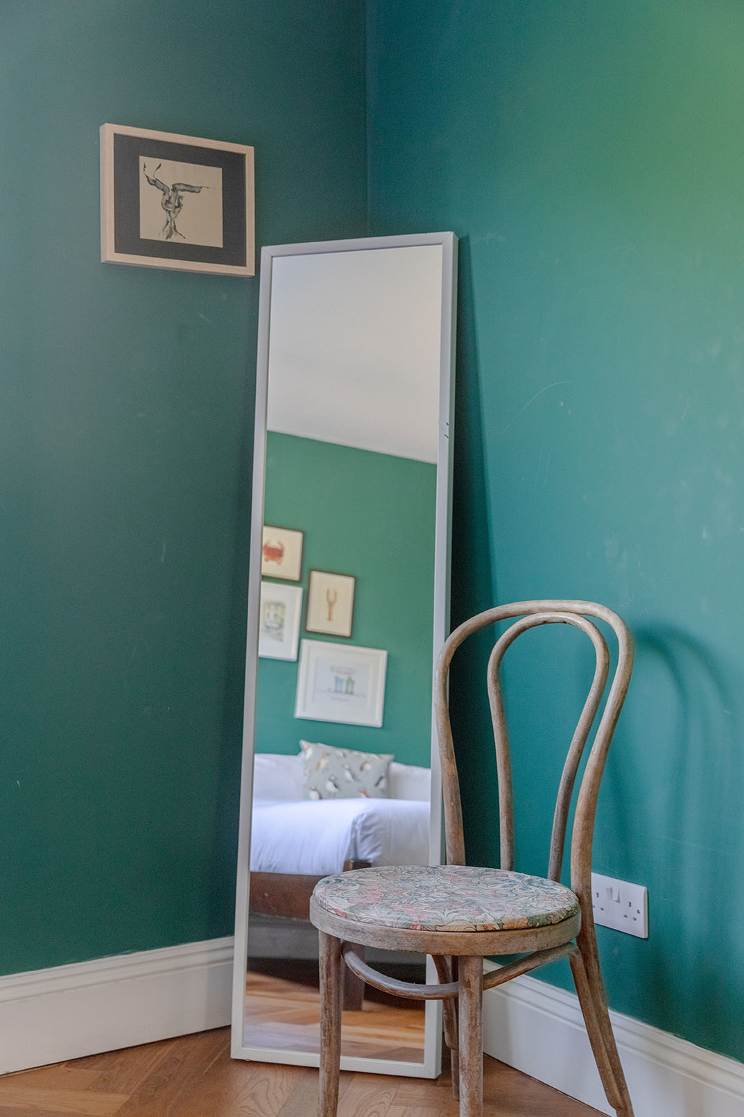 Corner of a bedroom with dark green walls, a full length mirror, an old wooden chair and the partial reflection of the bed with white sheets, captured for property photography as a detail for an airbnb listing