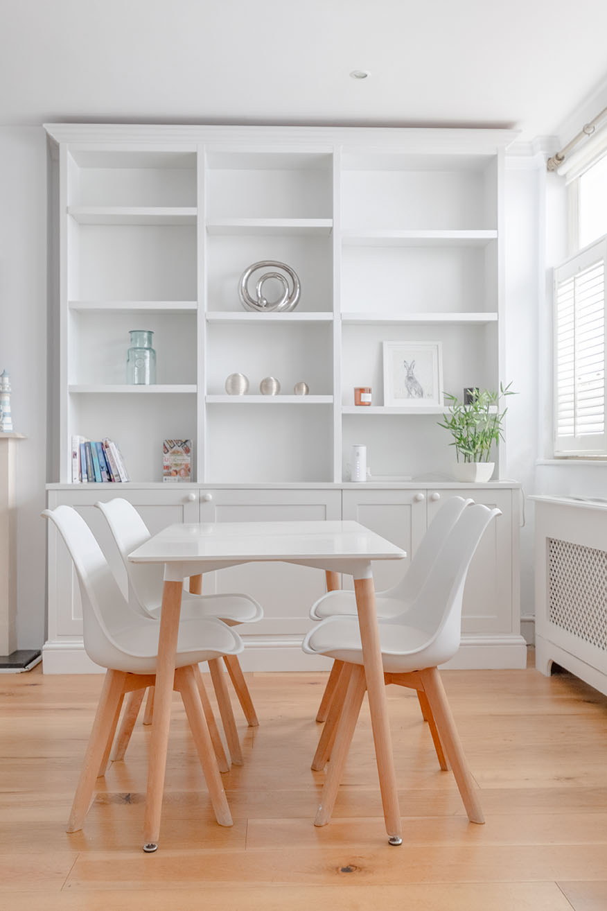 A dining room, white wooden dining table and chairs, a white bookshelf at the end of the room, bright sunny day.