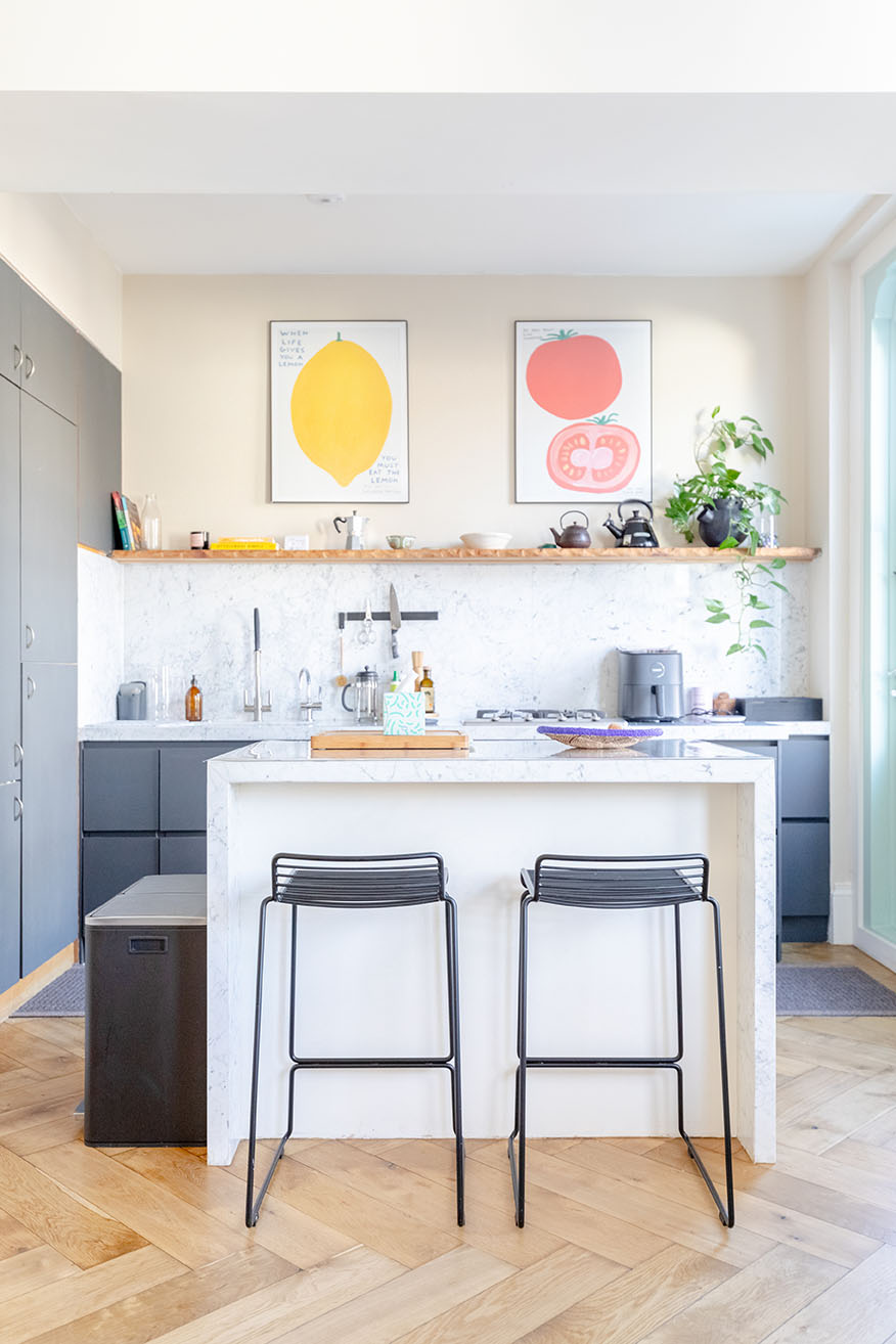 A vertical image of a kitchen with dark navy cupboards and a marble counter island with high stools in front of it.