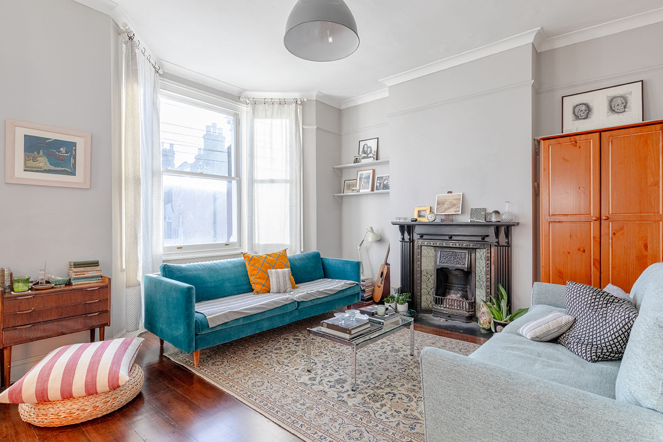 A warm decorated living room with an emerald green sofa, light grey walls, wooden floor, an old carpet, wooden cupboard and a fireplace, photographed for property photography.