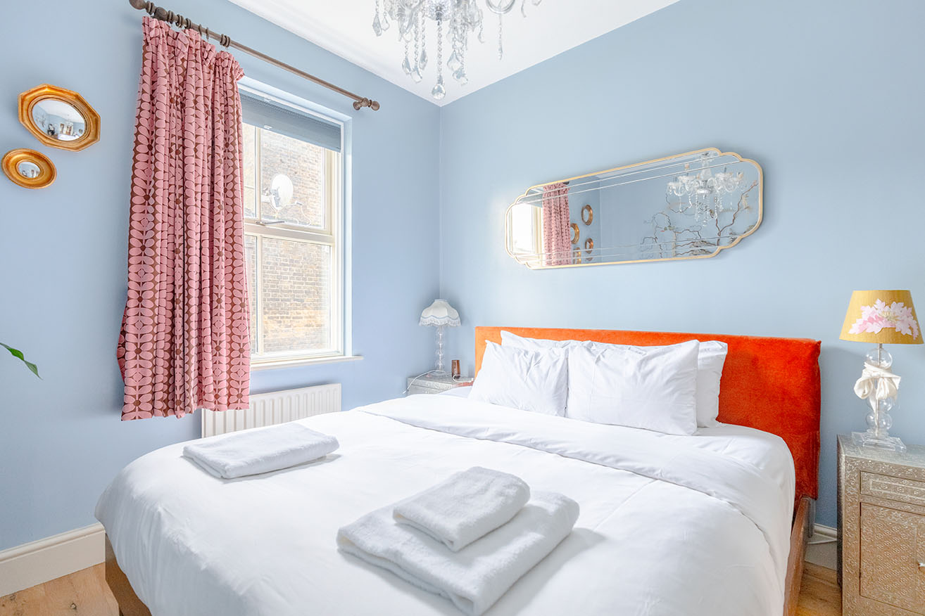 A small bedroom with light blue walls, a bright orange headboard of a bed with white linens, a large window, pink curtains and a mirror on the wall, cross angle.