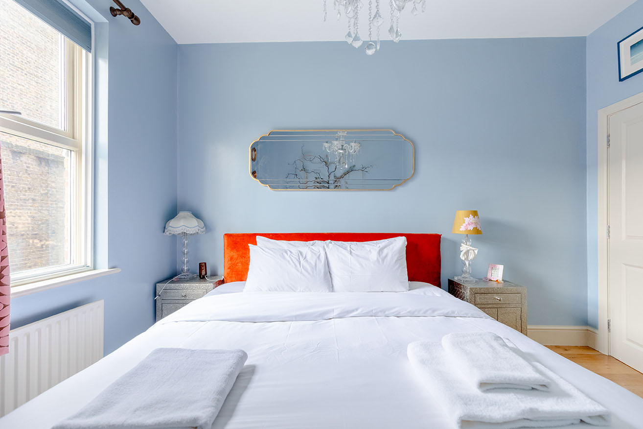 A small bedroom with light blue walls, a bright orange headboard of a bed with white linens, straight angle for property photography.