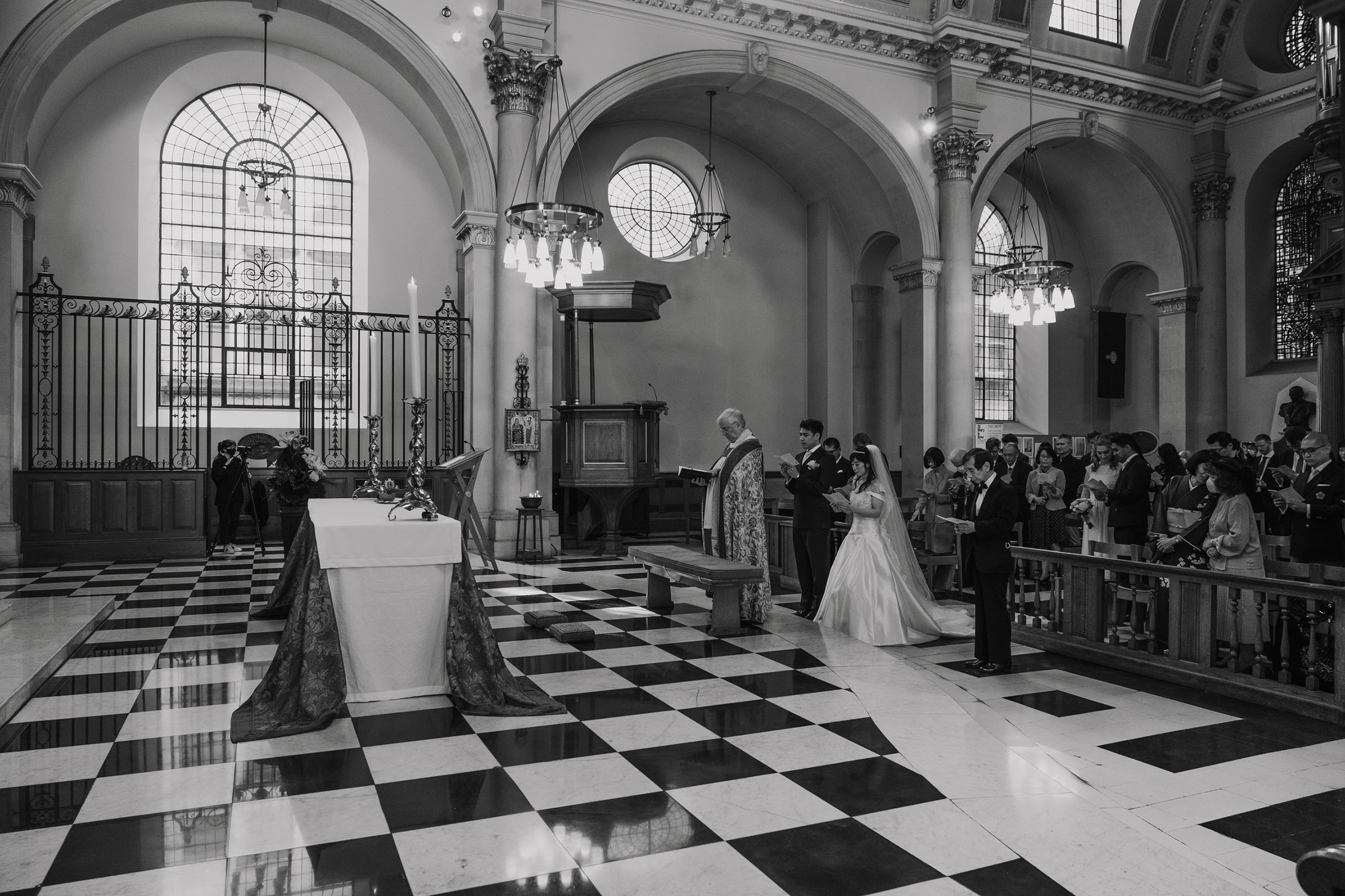 Inside a church in London, checkered floor, marrying couple with the priest, black and white wedding photography.