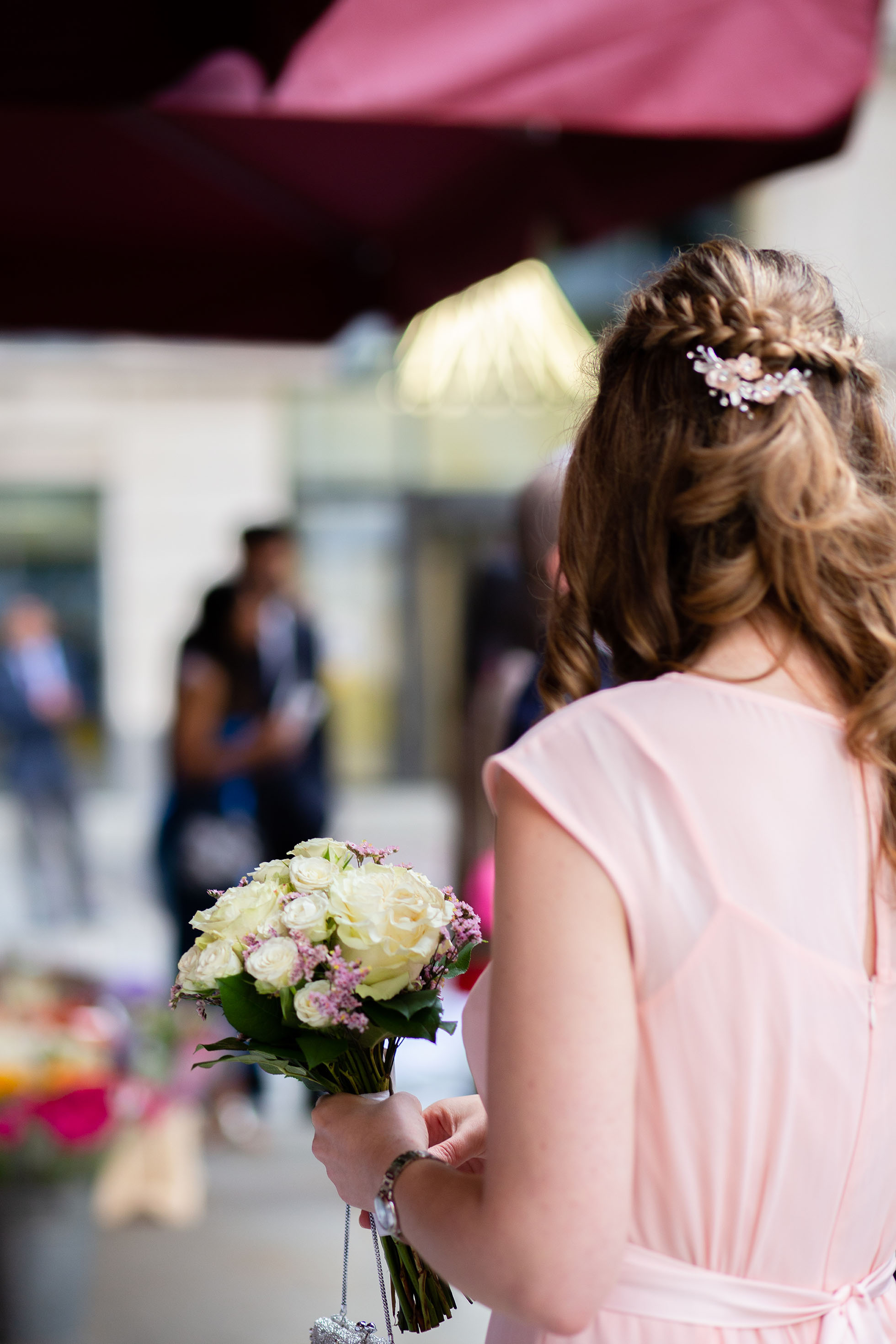 A bridesmaid holding the bridal bouquet, wearing a soft pink dress, captured from behind during wedding photography.